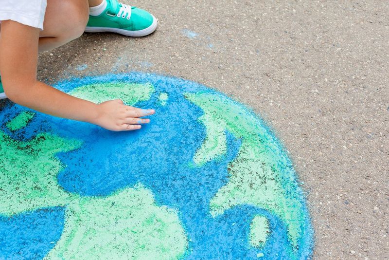 A partial view of a chalk picture of the globe in blue and green. To the left of the image a child’s right arm, right knee and left foot can be seen. They are rubbing the chalk with their finger.