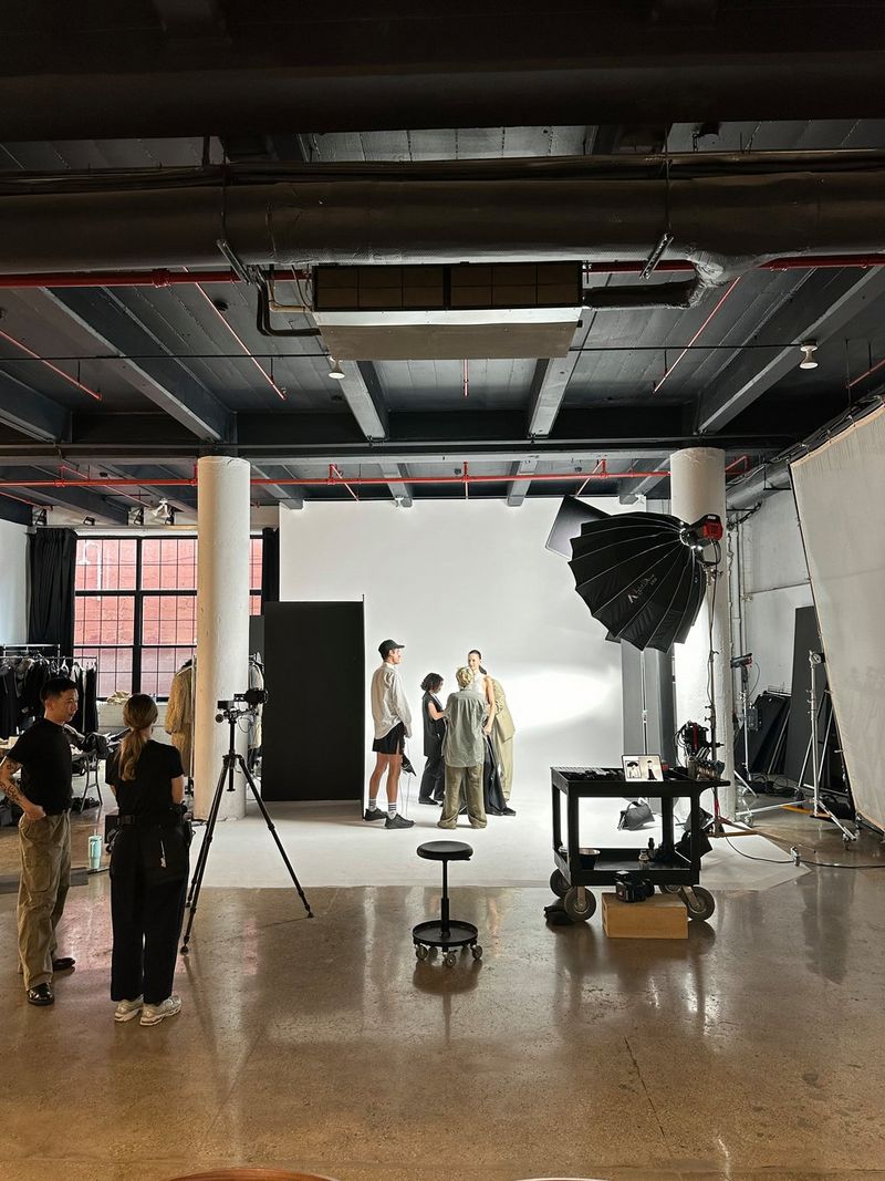A wide angle shot of a photo studio with three woman and a man standing on a white infinity curve. To the left, a man and woman operate a camera. 