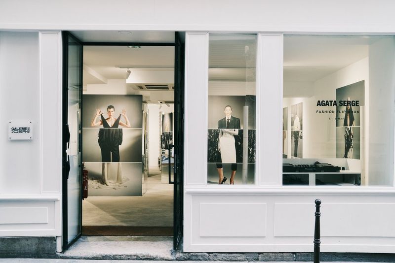 The exterior of an exhibition space in Paris with printed fashion portraits visible through the door and window. In front, a woman walks past. 