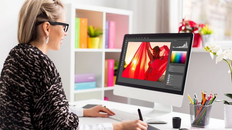 A bespectacled blonde woman sits at a white desk in a stylish office, She uses a graphics tablet to edit a photo of a figure in red on the screen.
