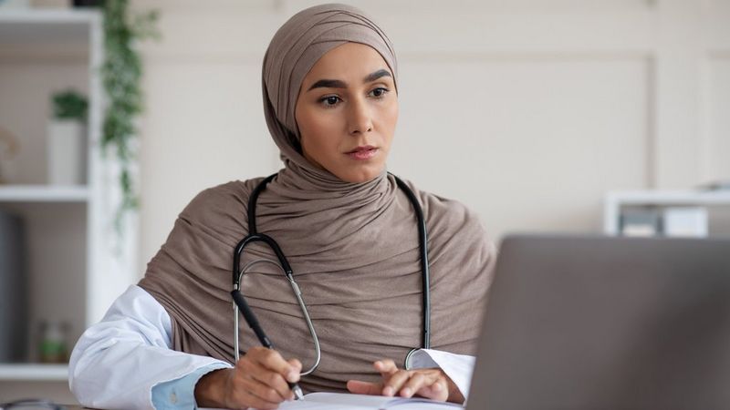 A female Muslim doctor sits at a laptop writing notes, she has a stethoscope around her neck.