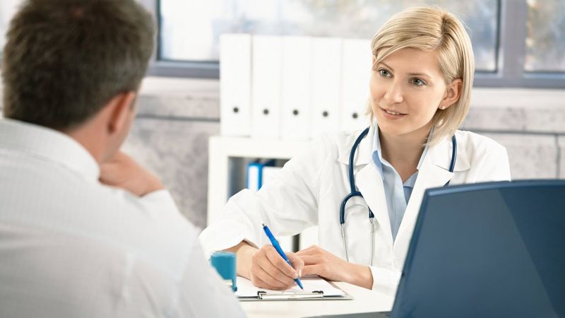 A female doctor sits at a laptop taking notes on a clipboard. She has a stethoscope around her neck. Opposite her with his back to the camera is a patient.