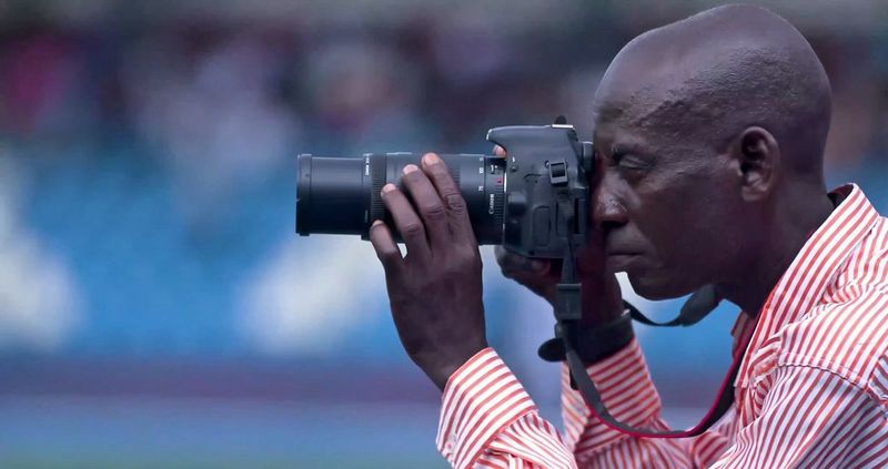 Akeem, wearing a red and white striped shirt, looks through the viewfinder of his new Canon camera. The background is a soft, blurry blue, likely a stadium.