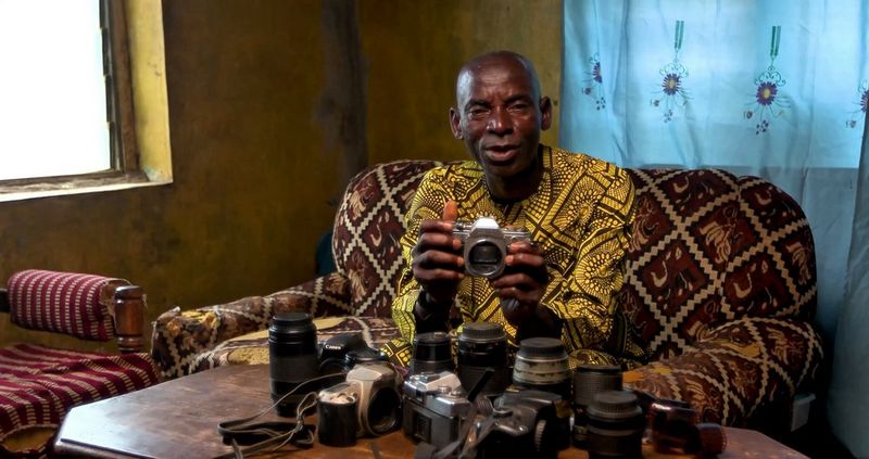 Akeem Olamide Olasun wears a vibrant yellow patterned shirt and sits on a patterned sofa, holding up a vintage Canon camera. Several other Canon cameras and lenses are spread across a wooden table in front of him.