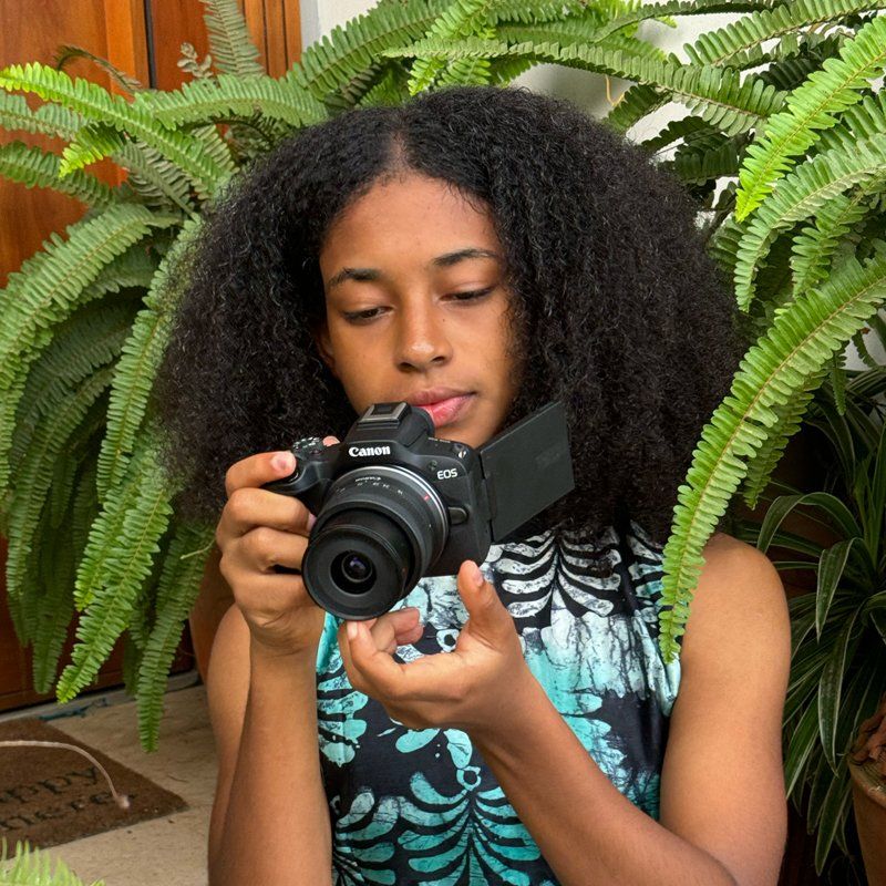 A young woman holds a Canon camera and looks into the rear screen. Behind her is a large green fern.