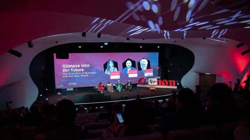 A stage, photographed from above and behind the audience. Four people sit on chairs on the stage beside a huge 3D Canon logo and a painting on an easel. Behind them is a screen showing photos of three of the people on the chairs. Next to their images are the words, “Glimpse into the future. The evolution of business in the new world.”