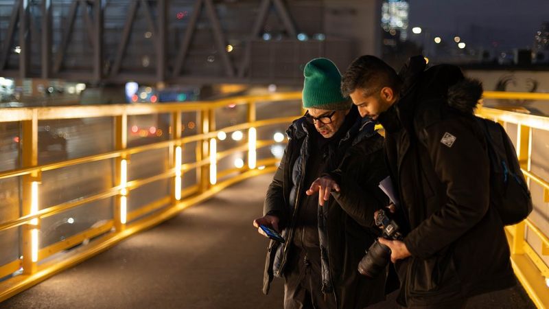 Aron and a contemporary stand on a lit pedestrian bridge at night, looking at a smartphone and holding a Canon camera.