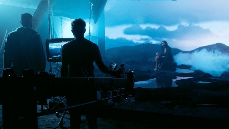 On a darkened film set, a woman sits in a misty, blue-lit artificial landscape. Two silhouetted crew members stand to one side, watching her on a screen.