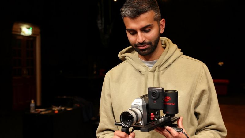 Aron Randhawa, bekleidet mit einem hellbraunen Kapuzenpulli, blickt hinunter auf seine Canon Super-8-Kamera, die mit verschiedenen individuell angepassten Aufsätzen ausgestattet ist.