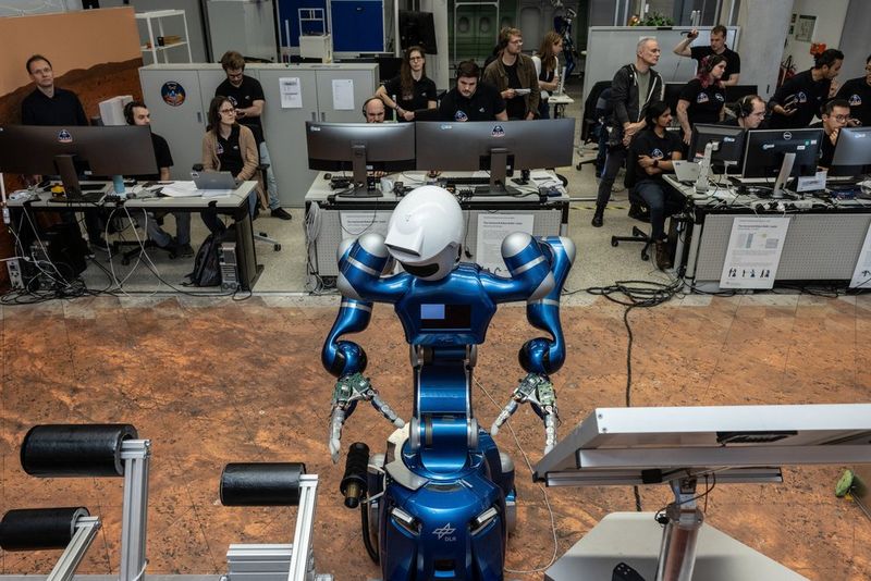 Een blauw-zilveren mensachtige robot staat met zijn rug naar een team van wetenschappers en ingenieurs, die hem blijkbaar bedienen vanaf hun bureaus met meerdere computerschermen.