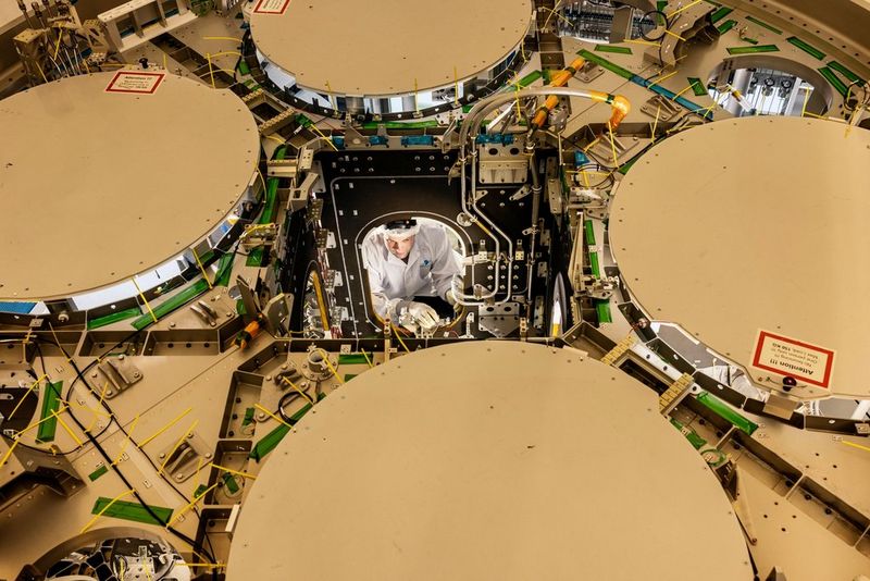 Op deze foto, genomen vanuit de lucht, is te zien hoe een technicus in een witte laboratoriumjas, met een haarnetje en beschermende handschoenen aan, in het centrale luik van het metalen frame van een ruimtevaartuig aan het werk is. Hij is zorgvuldig bezig met de interne bedrading en onderdelen.