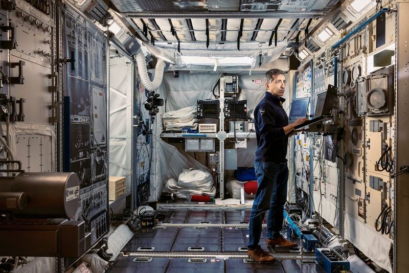 Een astronaut in vrijetijdskleding staat in een zeer gedetailleerde maquette van een ruimtestationmodule. Hij is bezig met een laptop die is bevestigd aan een wandpaneel vol ingewikkelde schakelaars, kabels en apparatuur.
