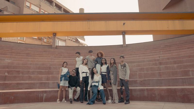Ten models pose in front of an amphitheatre-style set of steps. They are all wearing clothes from the Adaptive collection – jeans, shorts, belted shorts and utilitarian trousers and jackets. In the centre, one model sits in a wheelchair.