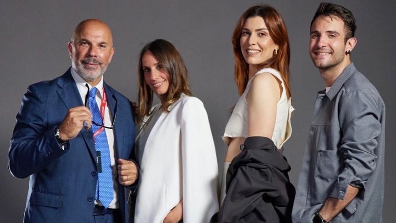 Four people stand in a row, smiling at the camera. On the left is a bearded man in a blue suit and tie. He holds his glasses in his right hand and his body is facing the camera. Next is a woman dressed in white with long brown hair, she stands facing him with her right side facing the camera. Also stood right shoulder to camera beside her are two further people – a taller red haired woman in a cropped, sleeveless cream top and black jacket, pushed off her back and gathered at her elbows. Then a man in a grey shirt.