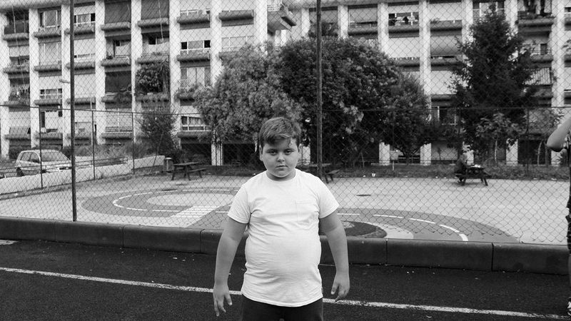 A black and white photo of a young boy standing in front of a playground, behind which are the apartments of the former Bastogi residence, looking directly at the camera with his hands by his sides.