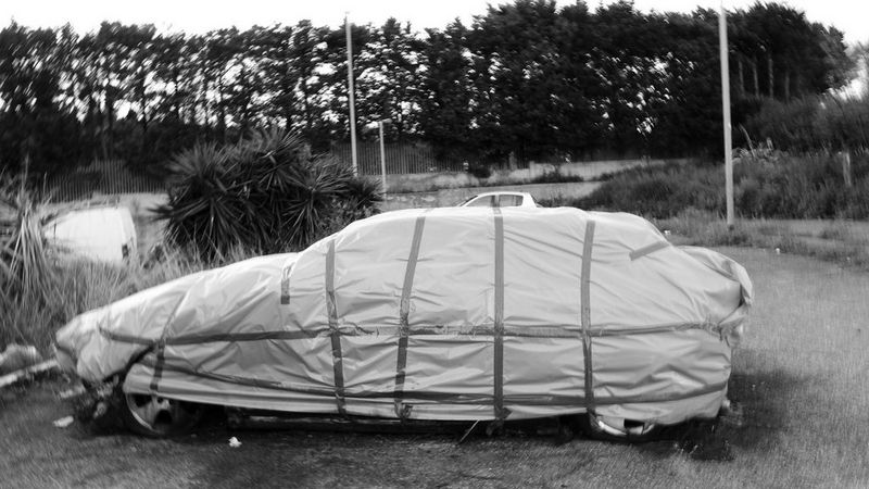 A black and white photo of a car covered in a taped-on tarpaulin. It is parked on a grassy patch of land, with tall bushes and trees in the background.
