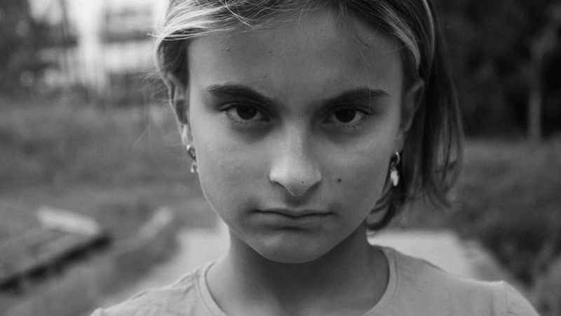 A black and white close-up photograph of a young girl’s face. She looks directly at the camera with a solemn expression.