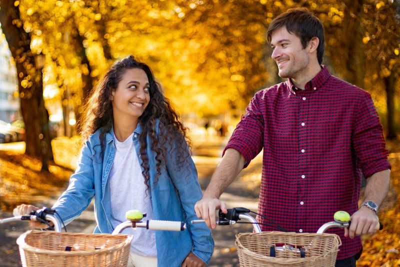 A young man and woman smile at each other as they push their bikes along a path with a backdrop of golden autumn leaves.