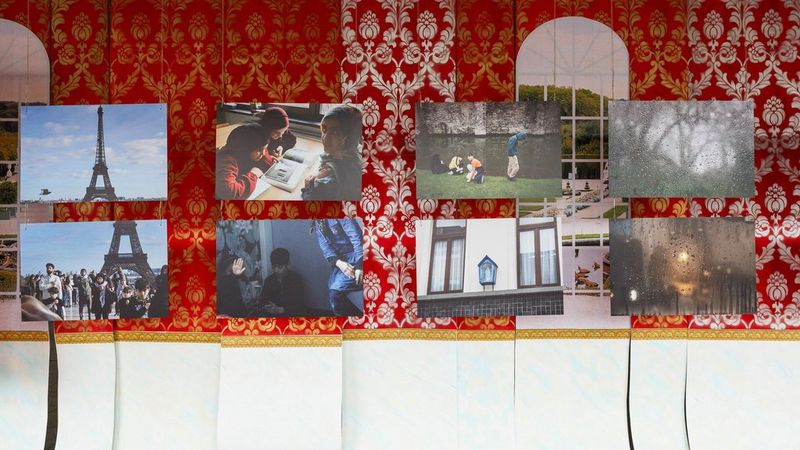 Eight printed images of various scenes and people hang on a red curtained wall.