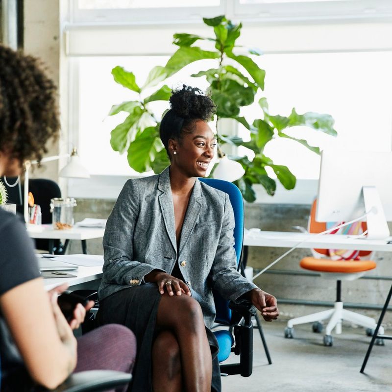 Woman sits on blue chair in front of a plant and talks to two people