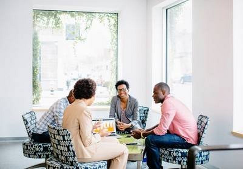 Two men and two women sit round a small table on comfy patterned black and white chairs looking at graphs on a laptop.