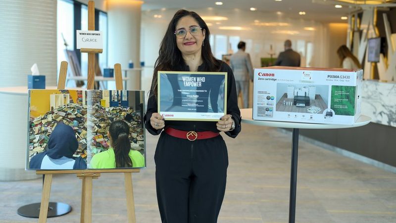 Grace Karim holds a certificate as she stands between an easel displaying the photograph of she and Somia looking at the destroyed books and a boxed Canon printer on a table.