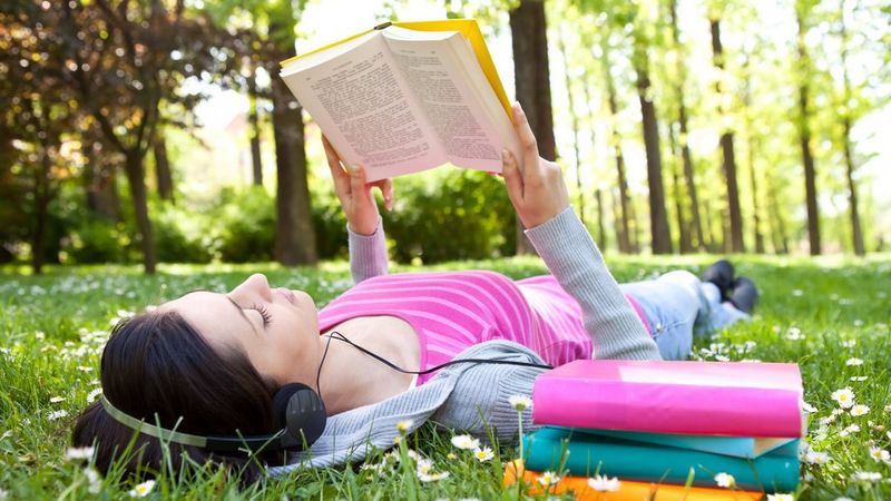 A young woman wearing headphones lies on the grass in a park, reading a book.