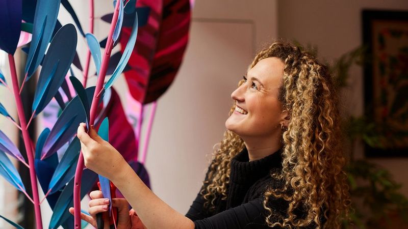 Artist Caroline Byrne smiles as she looks at the oversized blue and red paper flower sculptures that she holds. She has long blonde tightly curly hair and is wearing a black sweater.