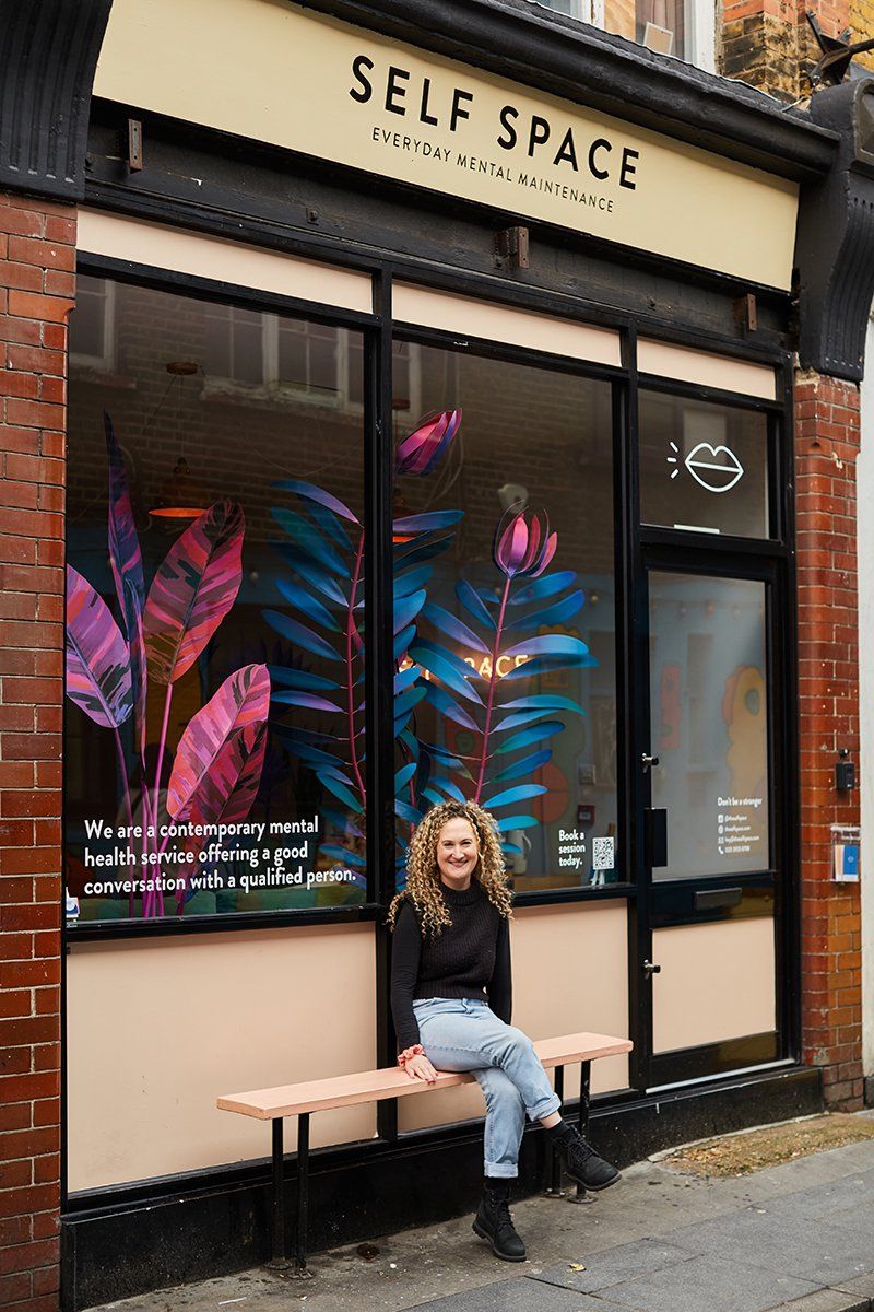Artist Caroline Byrne sits on a bench outside the ‘shop front’ of Self Space. Behind her in the window are her beautiful red and blue plant sculptures.