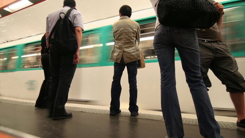 A photo, taken from ground angle, of five people stood on a subway platform. A white a green train speeds past them.