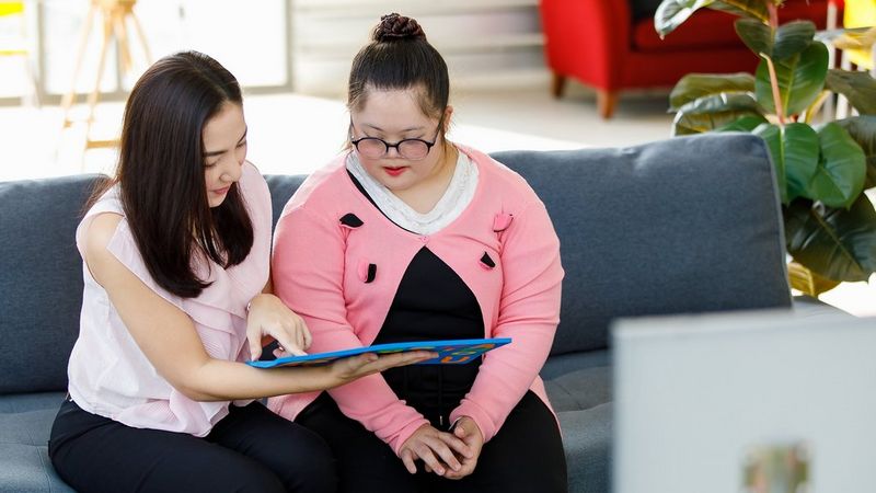 A female teacher sits on a grey couch with a student, holding a worksheet and pointing at it. She wears white sleeveless blouse and black trousers. Her hair is shoulder length and dark. The student, who sits to her right, is wearing a black dress with a white collar and a pink cardigan. She wears glasses and her hair is in a high bun. There is a green leafy plant to the right of the couch.