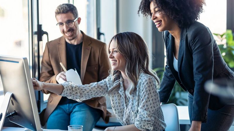 Three smiling people look at a computer monitor. A man in glasses, perching on a desk on the far left, a woman with blonde wavy hair and a white patterned blouse in the middle, pointing at the screen. On the right, a curly haired woman stands, leaning towards the screen. She wears a suit jacket over a white top and jeans.
