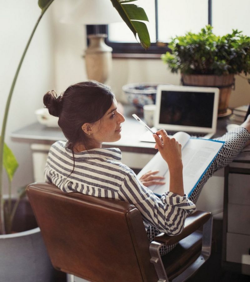 Women in stripy shirt sits in a brown leather chair while resting her legs on the desk beside a laptop as she reads a document.