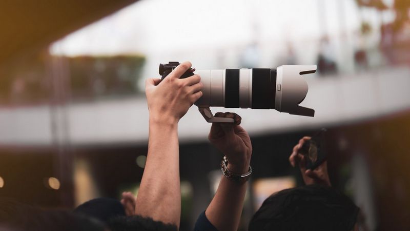 Hands holding a Canon mirrorless camera with a large white telephoto lens high above a crowd.