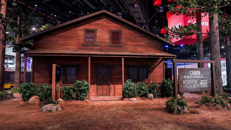 A brown wooden cabin, surrounded by dirt, rocks, trees and foliage, as though in the woods. Out the front of the cabin is a wooden swinging sign that reads “Canon”, “M.Night Shyamalan”, “Knock at the Cabin” and “Immersive movie experience”. To the top right is a huge suspended red cube with the Canon logo on all visible sides.
