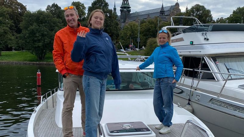 Camilla, her husband and one of her daughters stand on their moored family boat, smiling. There is a church-like building and many trees in the background.