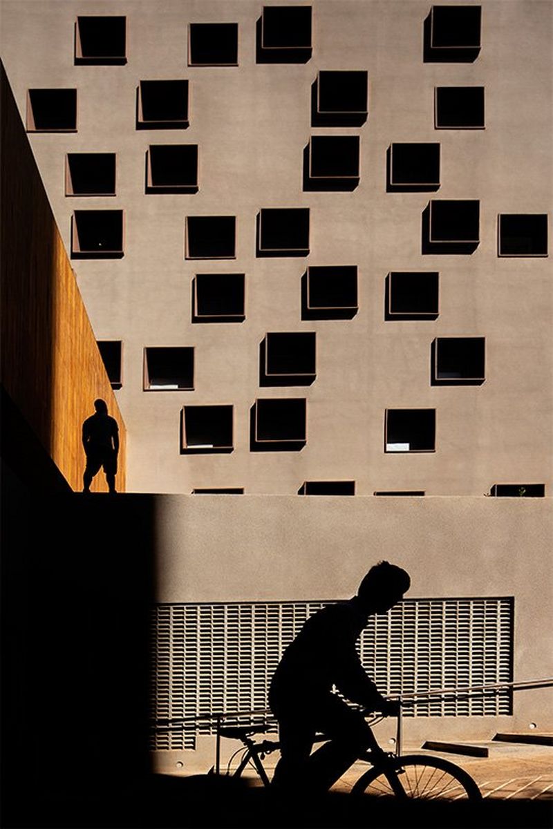 A silhouette of a cyclist passing a large concrete building with many windows. Photo by Fernando Guerra with a Canon TS-E 50mm f/2.8L MACRO lens.