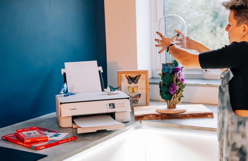 Eine Frau setzt eine Glaskuppel über ein Schmetterlingsdisplay, das neben einem Canon Drucker und einer Packung Canon Papier steht.