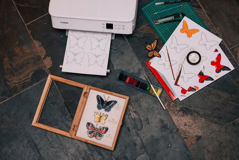 Ein Canon Drucker, ein offener Rahmen mit drei Papierschmetterlingen und Papierbögen mit Schmetterlingskonturen auf einer Arbeitsfläche.
