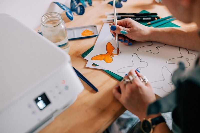 Eine Frau malt eine Schmetterlingsvorlage leuchtend orange aus. Auf dem Tisch neben ihr steht ein Canon Drucker.