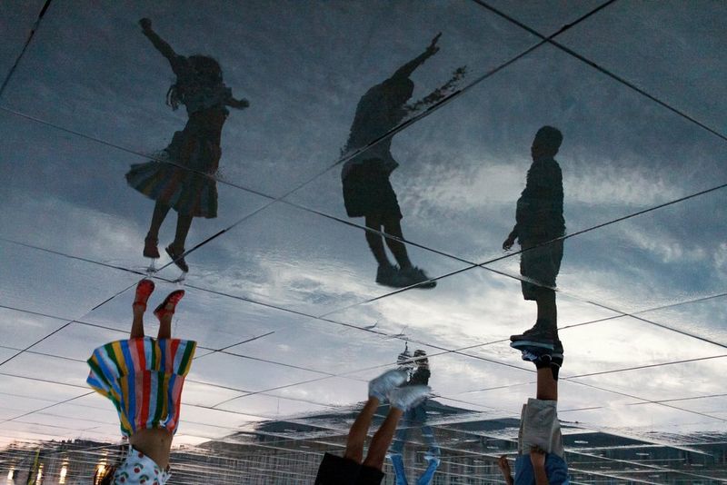 Vista invertida de unos reflejos sombríos de tres niños saltando sobre una zona pavimentada mojada. Los reflejos y el cielo sobre ellos parecen flotar por encima de la escena real, que solo es visible en la parte inferior de la imagen.