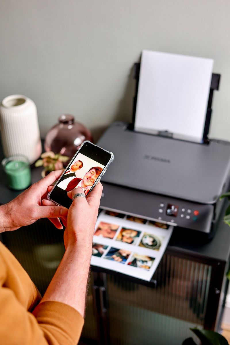 A pair of hands holds a smartphone with a photo on the screen in front of a Canon PIXMA printer with a sheet of photos in the process of being printed.