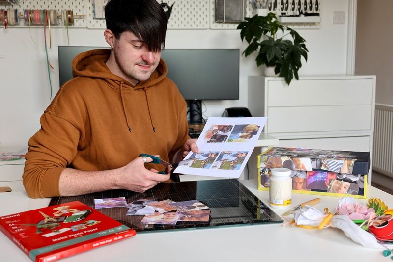 A young man uses scissors to cut around photographs printed on a Canon PIXMA printer. On the table beside him is a pack of Canon paper, glue and a part-finished découpage shoe box.