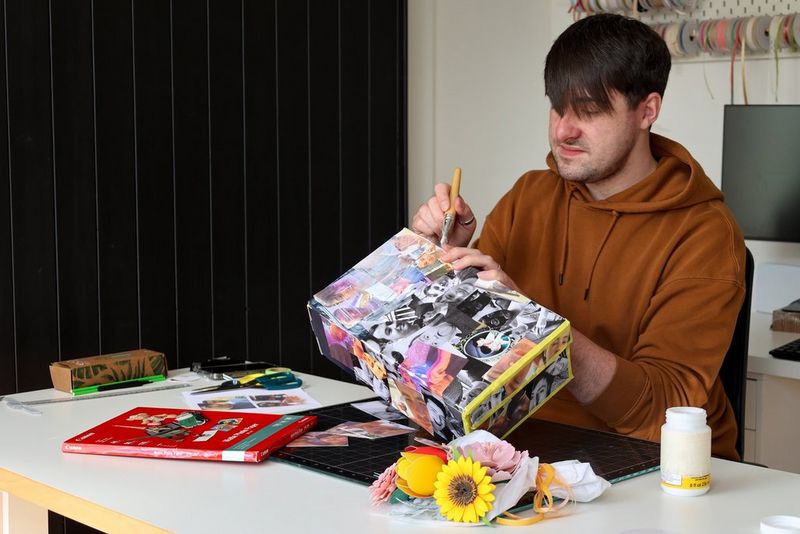 A young man uses a brush to glue down the finishing touches to a découpage gift box with photos on. A papercraft flower bouquet, pack of Canon paper and crafting tools are also on the table.