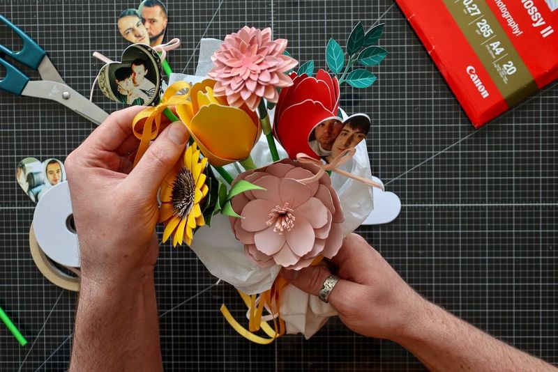 A pair of hands adds sticks with personalised heart-shaped photos to a papercraft flower bouquet.