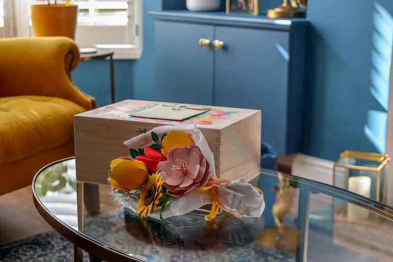 A papercraft bouquet sits on a table in front of a wooden box with a card on top in the middle of a living room.