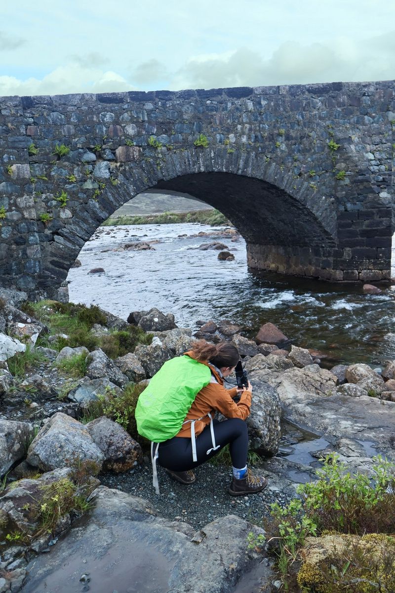 Vlogger Niamh Mackinnon crouches on rocks by a stream under a stone bridge, filming a video with a Canon EOS R50 V.