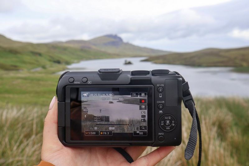 Vlogger Niamh Mackinnon holds a Canon EOS R50 V, with the rear screen showing the scene she is filming, a calm Scottish loch with a steep mountain rising in the background.
