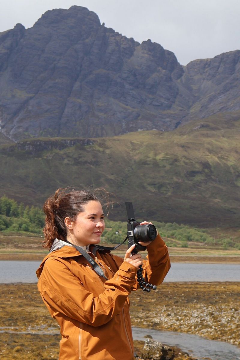  Vlogger Niamh Mackinnon stands near a loch on the Isle of Skye with mountains rising behind and films a video with the Canon EOS R50 V.