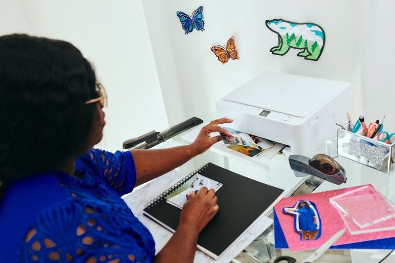 Kunsthåndværkeren Juliet Amma tager et fotoprint fra en Canon PIXMA-printers outputbakke med en bunke print, farvet papir og andre hobbyartikler på bordet foran hende.